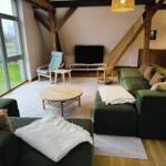 Wohnzimmer mit grüner Sofa, Holztisch und Fernseher. Große Fenster und Holzbalken.