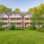 Rotes Mehrfamilienhaus mit weißen Balkonen und grünem Garten vor Bäumen.