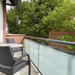 Terrasse mit Tisch und zwei Stühlen, Glasgeländer und grüner Hintergrund.