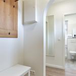 Flur mit Holzregal und weißen Möbeln, Blick in das Badezimmer mit Toilette.