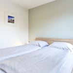 Ein Bett mit weißen Laken und Kissen, daneben Nachttisch mit Lampe und Wandbild.