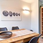 Esszimmer mit Holztisch, Stühlen und Wandbild mit Steinen. Bücherregal mit Pflanze und Glas.