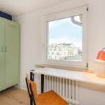 Ein Arbeitszimmer mit Schreibtisch, Stuhl und grünem Schrank. Fensterblick auf Gebäude.