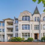 Villa Mailin mit markanter Fassade und Turm. Grüne Bäume und Beet umgeben das Gebäude.