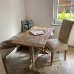 Holztisch mit Stuhl und Sitzbank an Fenster, gemusterte Fliesen und Kissen mit Ankermotiv.