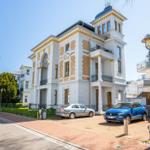 Historisches Gebäude mit weißen Wänden, gelben Akzenten und Balkonen. Zwei Autos parken vor dem Haus.