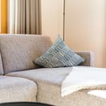 Ein grauer Sofa mit Kissen und Decke vor Fenster mit Vorhängen.