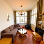 Wohnzimmer mit Sofa, Glas-Tisch und Fernseher. Große Fenster und Kronleuchter.