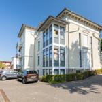 Haus mit mehreren Etagen, großen Fenstern und gepflastertem Hof mit parkenden Autos.