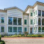 Villa Perkunos mit grünen Fensterrahmen und Balkonen, umgeben von Garten und Zaun.