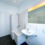 Badezimmer mit Dusche, Waschbecken und Spiegel. Fliesenwand und dunkler Boden.