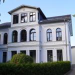 Ein weißes, dreigeschossiges Haus mit Bogenfenstern und einem Balkon. Umgeben von grünen Sträuchern.