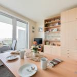 Essbereich mit Tisch, Stühlen und Blick auf Balkon. Hintergrund: Wohnbereich mit TV und Schrank.
