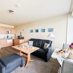 Wohnbereich mit Sofa, Esstisch und Küchenzeile. Große Fenster, Bilder an der Wand.