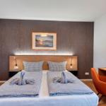 Doppelbett mit Kissen und Handtüchern, Schreibtisch mit Laptop und orange Stuhl.