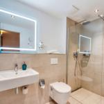 Badezimmer mit Dusche, Waschbecken und Toilette. Spiegel mit Beleuchtung und Handtuch.