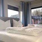 Das Bett mit weißen Laken und Kissen. Durch Fenster ist Außenansicht mit Häusern und Bäumen sichtbar.