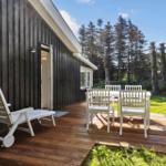Deck with white table and chairs, lounge chair, and wooden flooring beside a house with dark siding.