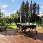 Deck with table and chairs, surrounded by greenery and trees. House with window on the right.