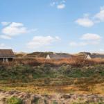 Häuser in der Ferne, umgeben von Dünen und Grasland unter blauem Himmel mit weißen Wolken.