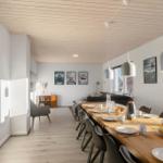 Dining room with long wooden table and chairs, living area in background, windows with view outside.