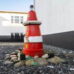 A red plastic lighthouse stands on stones in front of a house.