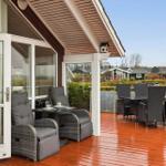 Deck with seating area and dining table. Wooden planks and glass railing. View of residential houses.