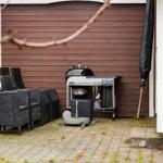 Outdoor area with grill, seating group, and umbrella. Paved stone floor and wooden wall in background.