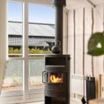 A wood stove with a kettle sits before large windows overlooking rooftops.