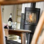 A wood-burning stove stands in a room with wooden walls and a table with a bottle and glasses.