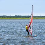 A windsurfer glides across calm waters with green land in the background.