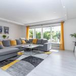 Wohnzimmer mit grauem Sofa, gelben Kissen und großem Fenster. Teppich und Wohnmöbel sind modern.