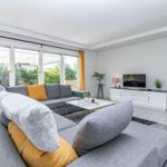 Wohnzimmer mit grauem Sofa, gelben Kissen und großem Fenster zur Terrasse.
