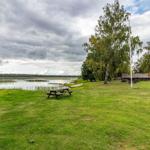 Grüner Rasenbereich am See mit Picknicktischen und Boot. Hintergrund: Wald und bewölkter Himmel.