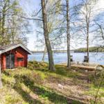 Rotes Holzhaus am See mit Terrasse und Stühlen zwischen Bäumen.