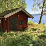 Rotes Holzhaus am See mit grüner Umgebung und einem kleinen Tisch vor der Tür.