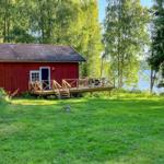 Rotes Holzhaus mit Terrasse am See, umgeben von Bäumen und Gras.