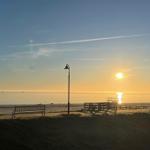Sonnenuntergang am Strand mit Bank und Leuchtturm im Hintergrund.