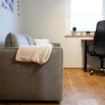 Ein grauer Sofa mit Decke, Schreibtisch und Stuhl im Raum.