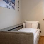 Ein Bett mit weißer Bettwäsche und Kissen. Fenster mit Jalousie und Pflanzen.