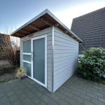 Ein kleines, graues Gartenhaus mit Glasfront und Dachvorsprung auf gepflastertem Boden.