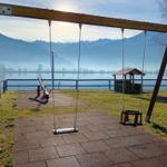 Kinderspielplatz mit Schaukel und Rutsche am See mit Bergen im Hintergrund.