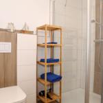 Badezimmer mit Dusche, Holzregal und Toilette. Blaue Handtücher und Deko auf Regal.