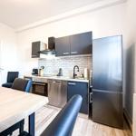 Küche mit Edelstahlgeräten, Holztisch und schwarzen Stühlen. Hintergrund: Backsteinwand und Kühlschrank.