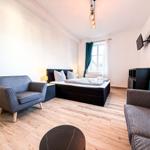 Schlafzimmer mit Bett, Sessel und TV. Holzboden und Fenster mit Vorhängen.