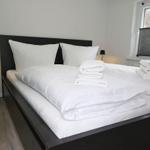 Ein Bett mit weißer Bettwäsche und gefalteten Handtüchern. Links steht ein Nachttisch mit Lampe.