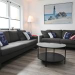 Zwei graue Sofas mit Kissen stehen um einen runden Tisch herum. An der Wand hängt ein Leuchtturm-Bild.