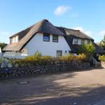 Haus mit strohgedecktem Dach, weiß verputzt, umgeben von Steinmauer und Bäumen.