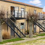 Two-story building with balcony and metal staircase. Trees and paved area.