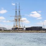 Historic sailing ship in harbor with modern buildings in background.